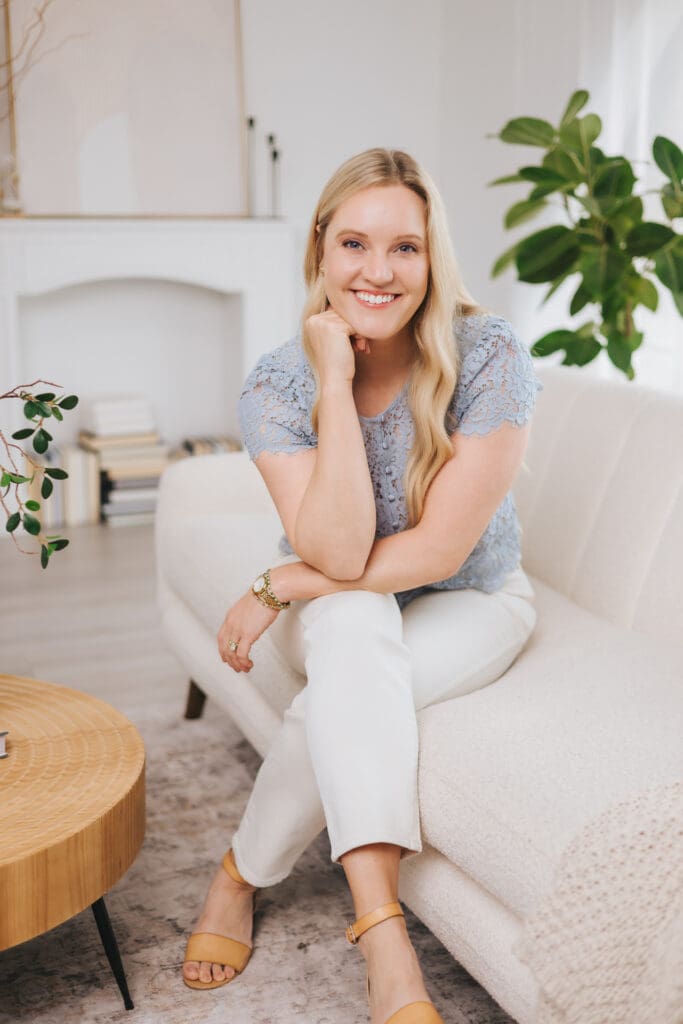 Jamie Urban wears a blue lace shirt and white pants sitting on a white couch staring ahead.