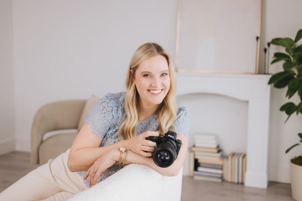 Jamie Urban sits on a white couch leaning over the back with one arm while the other arm holds her Nikon camera.