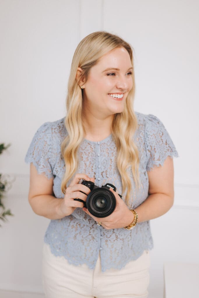 Jamie Urban wears a blue lace shirt, holds her Nikon camera and laughs looking off to her left.