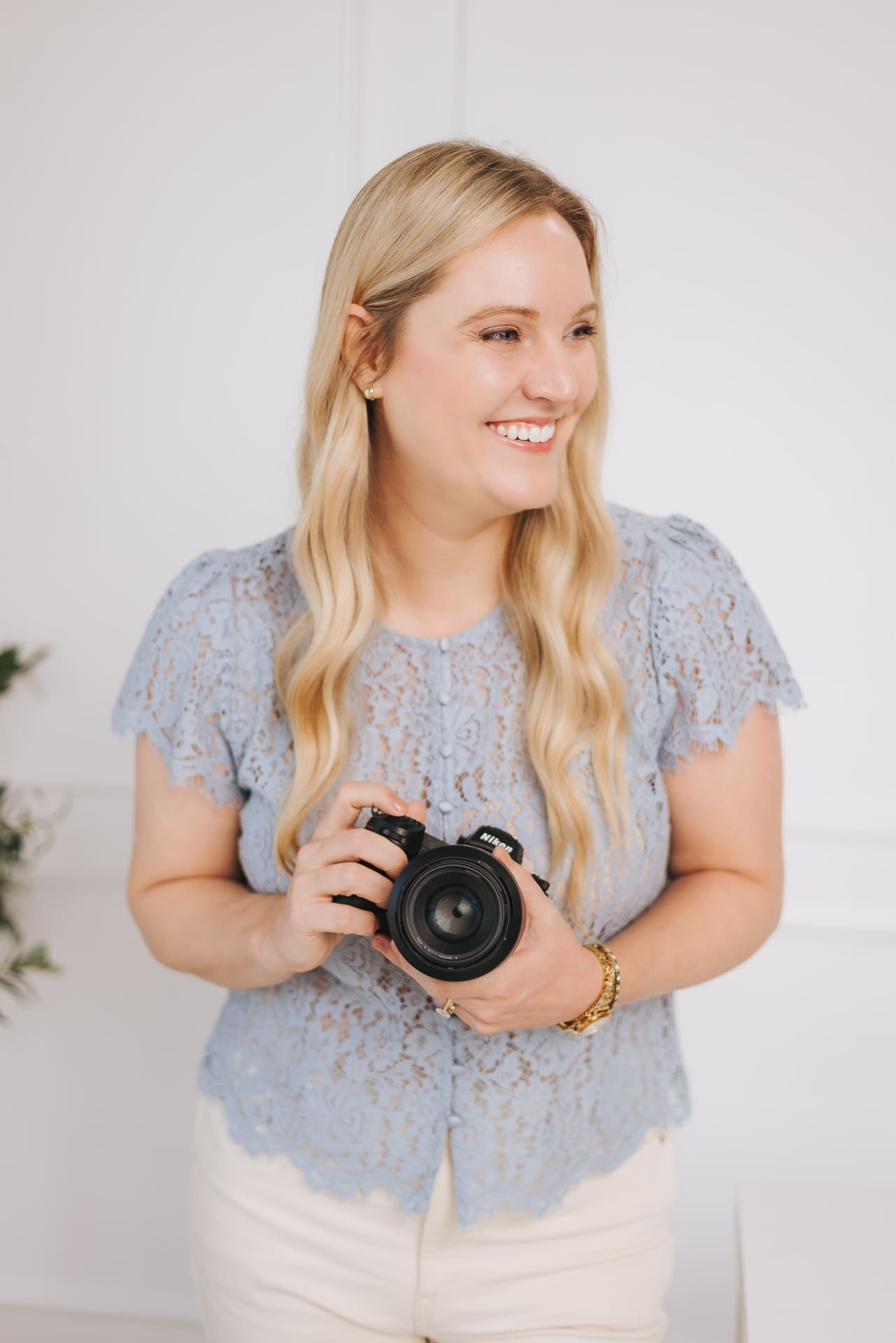 Jamie Urban stands against a white wall holding her Nikon camera smiling looking off to the left.