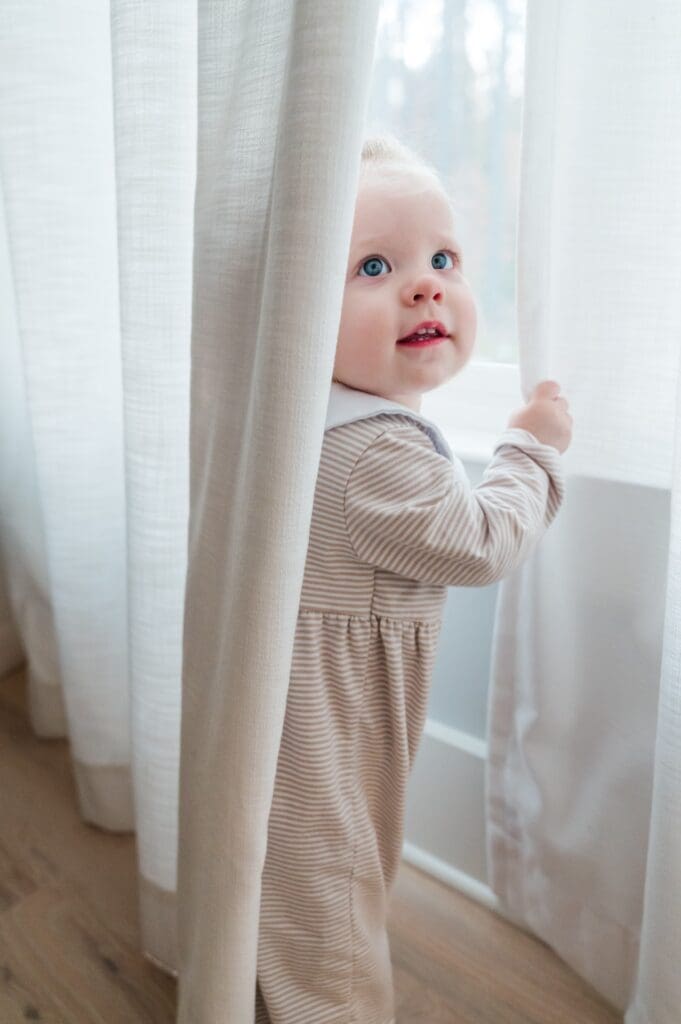 Small caucasian baby in beige striped onesie stands peeking out from behind white curtain.
