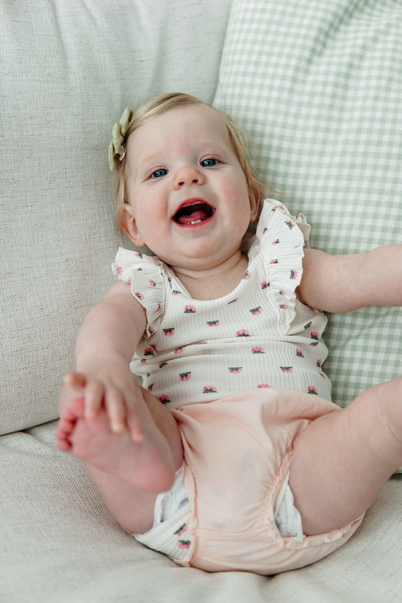 Small caucasian baby in white onesie with pink flowers sits on beige couch with green gingham pillow. Baby has open mouth smile while grabbing toes.