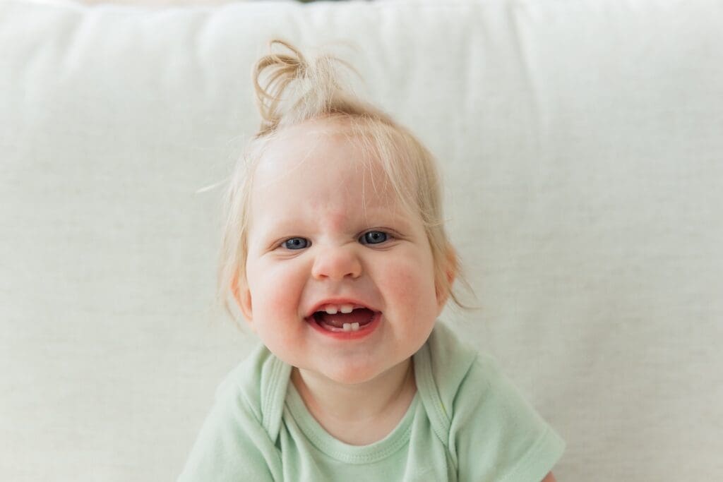 Small caucasian baby in green onesie sits on beige couch leaning forward. Baby has a top pony tail and a toothy open mouth smile.