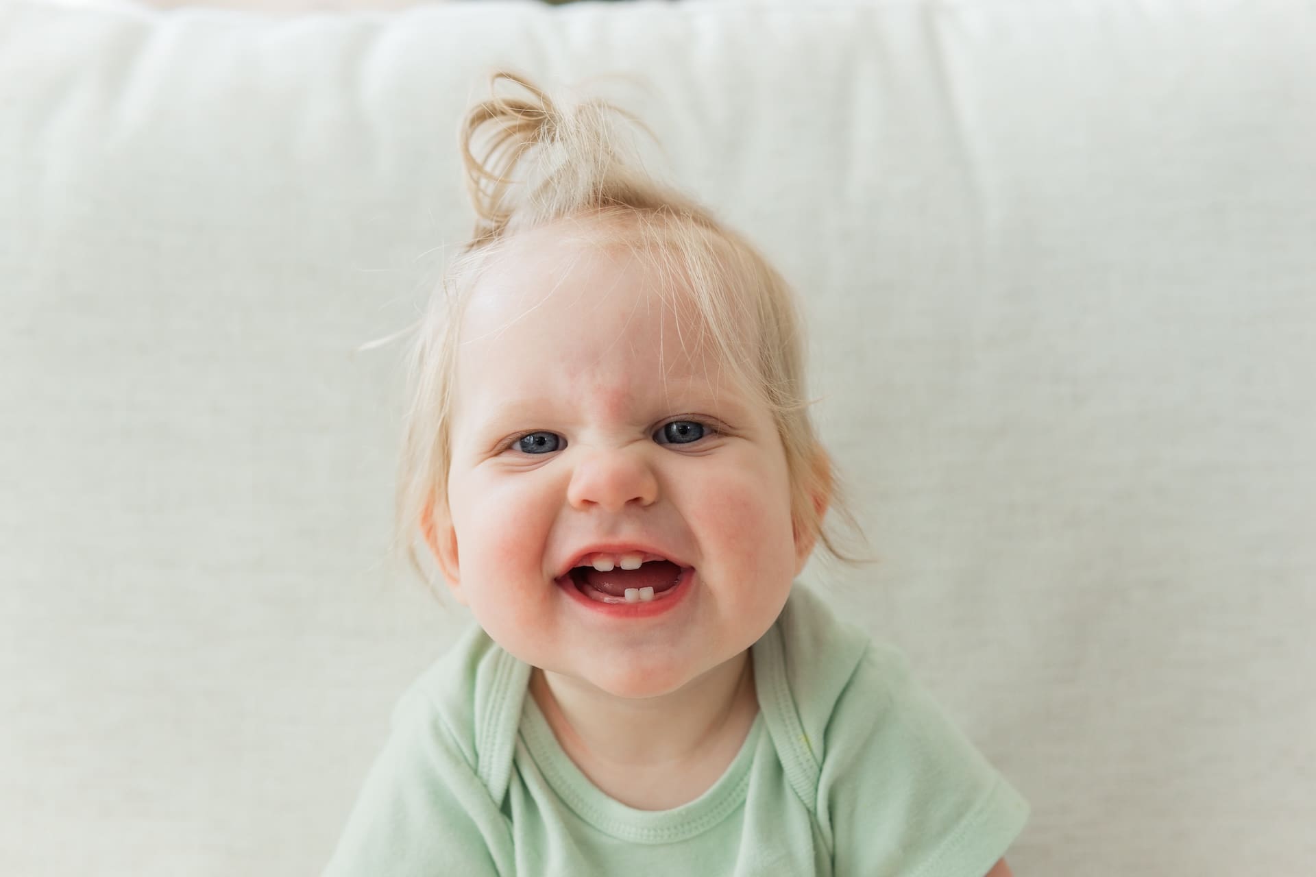 Baby with green onesie and pony tail sits on beige couch with a scrunchy nose, toothy smile.