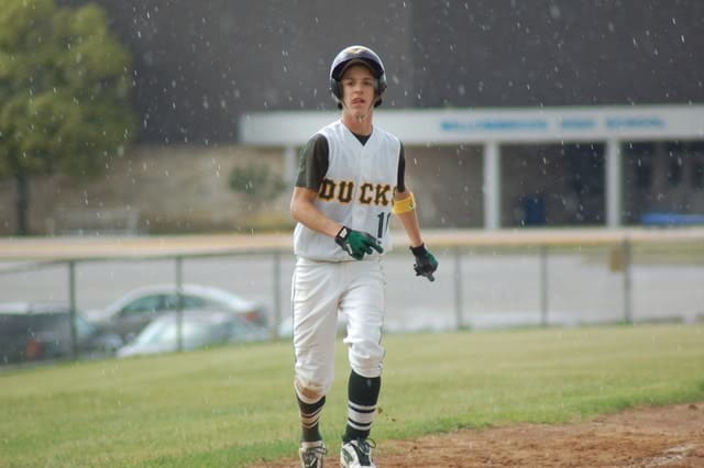High school aged caucasian boy in white baseball jersey with number ten runs in the rain down the 3rd base line.