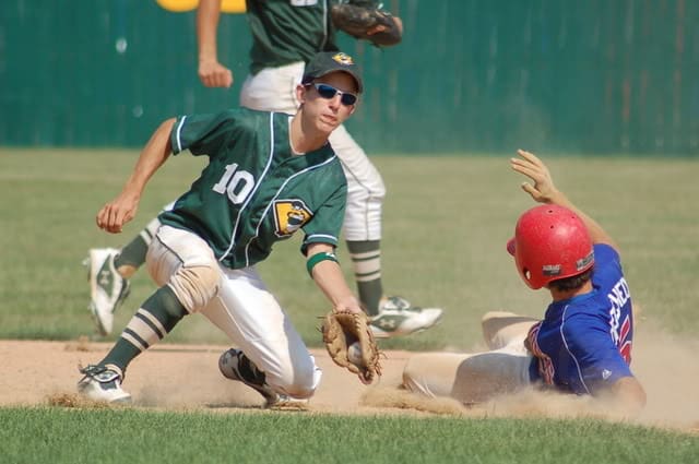 High school aged caucasian boy in green baseball jersey with number ten catches baseball to tag a runner sliding into the base. Runner is wearing a royal blue jersey and red helmet.