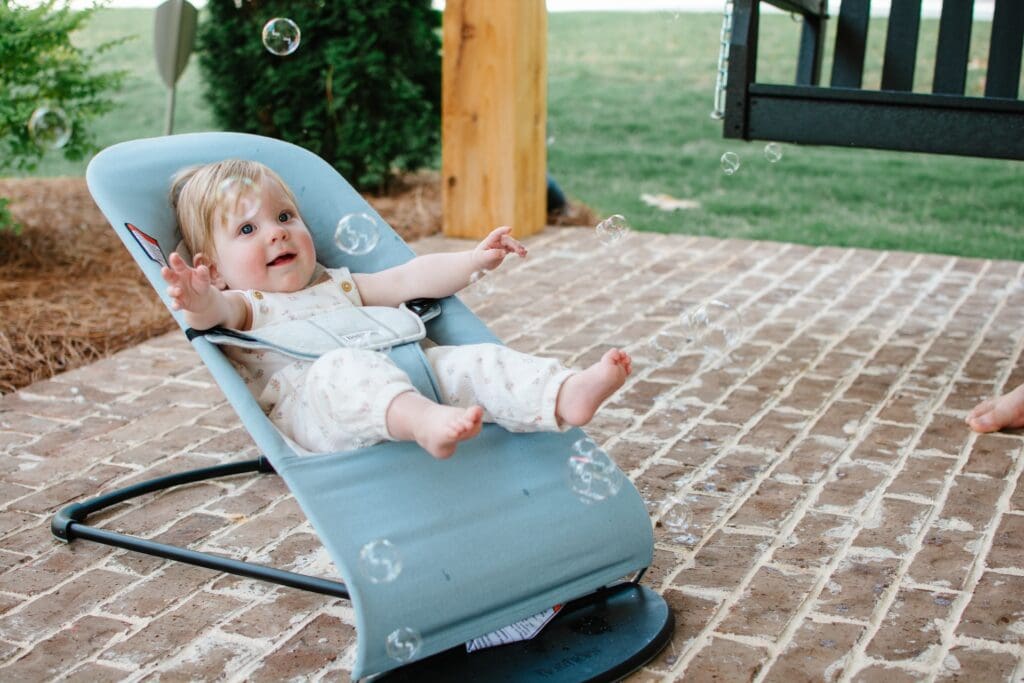 Small baby in a white floral jumpsuit sits in blue babybjorn bouncer on brick patio while bubbles fly overhead through the air.