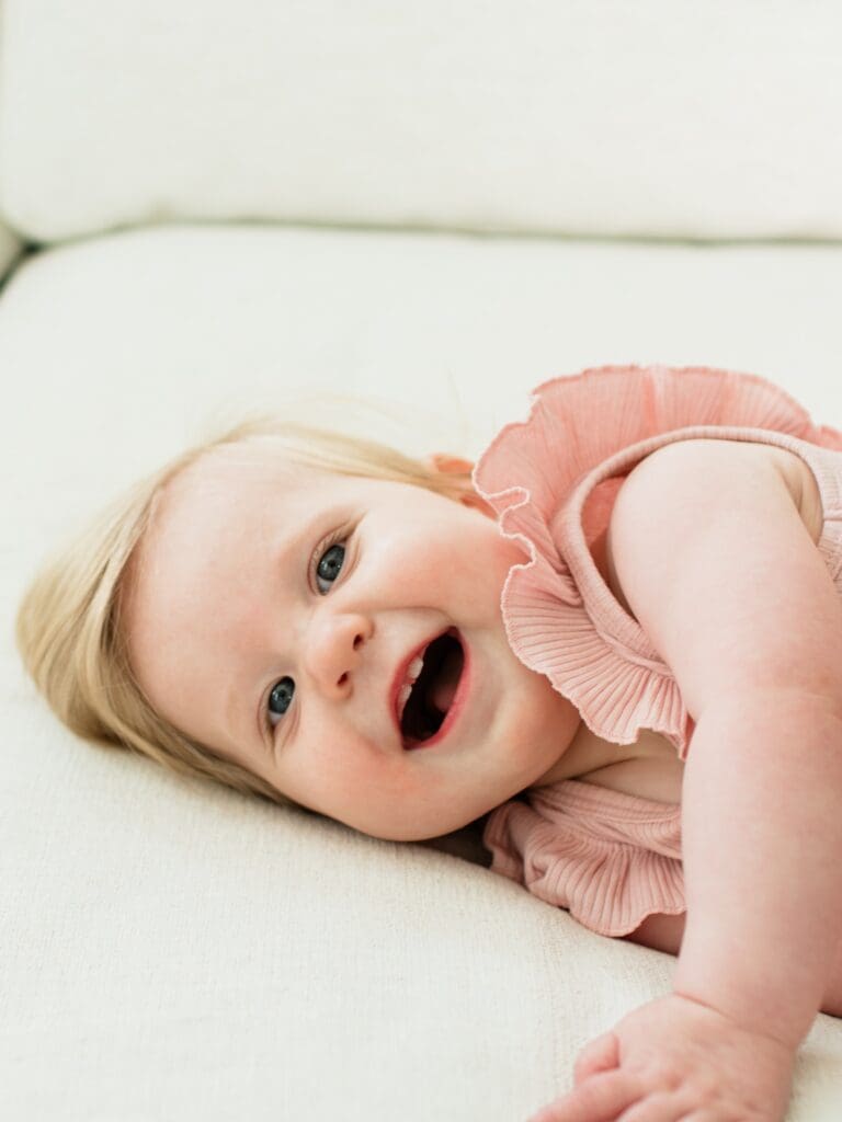 Small caucasian baby in pink onesie lays horizontally on beige couch. Baby has a toothy open mouth smile.