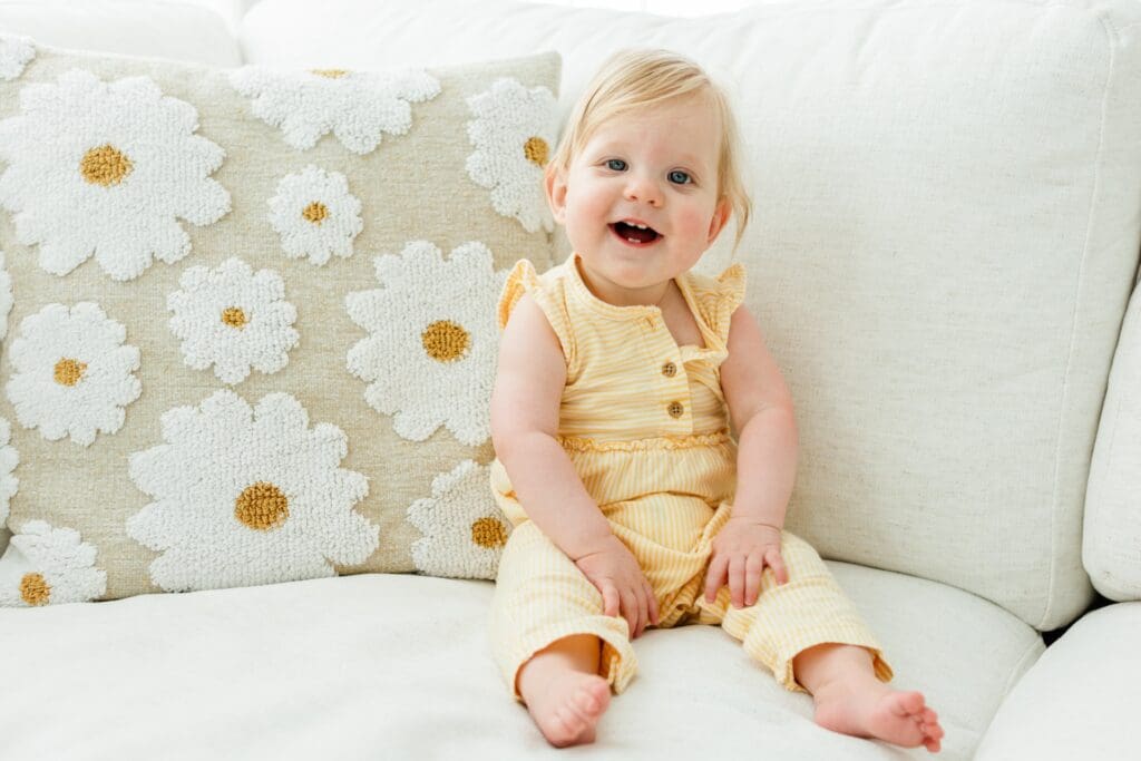 Small caucasian baby in yellow striped jumpsuit sits on beige couch. Baby has a toothy open mouth smile.
