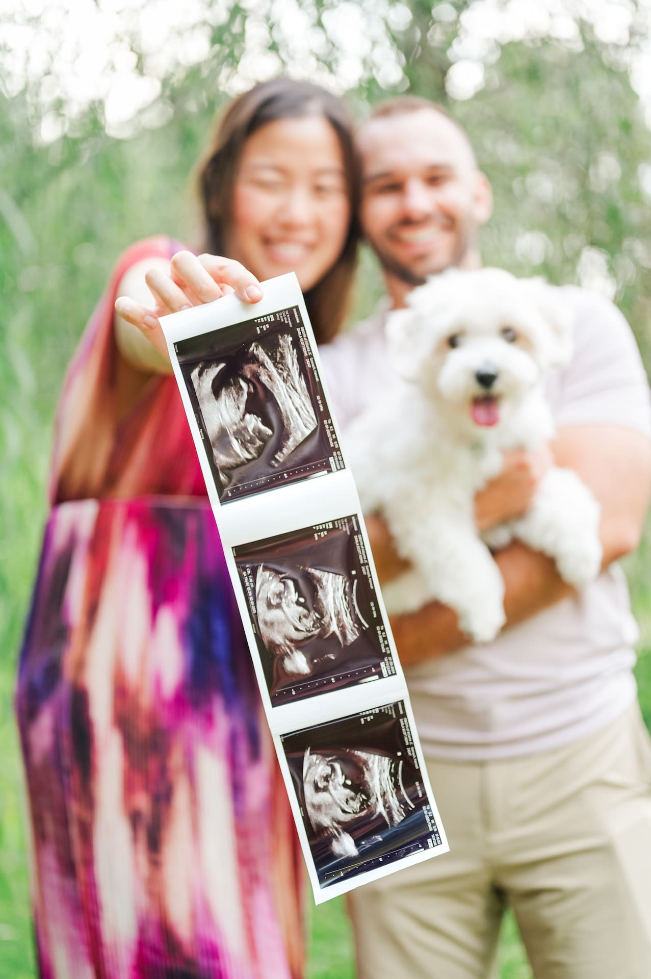 Young Korean woman stands next to young caucasian man holding small white dog while holding ultrasound pictures outdoors amongst greenery.