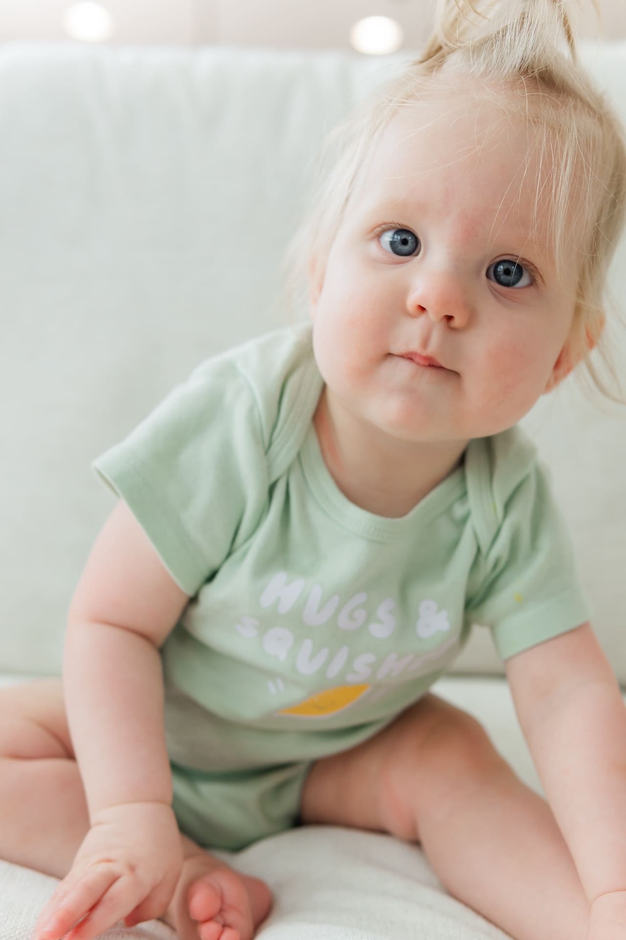 Baby in green onesie that says "hugs & squishes" sits leaning forward on a beige couch.