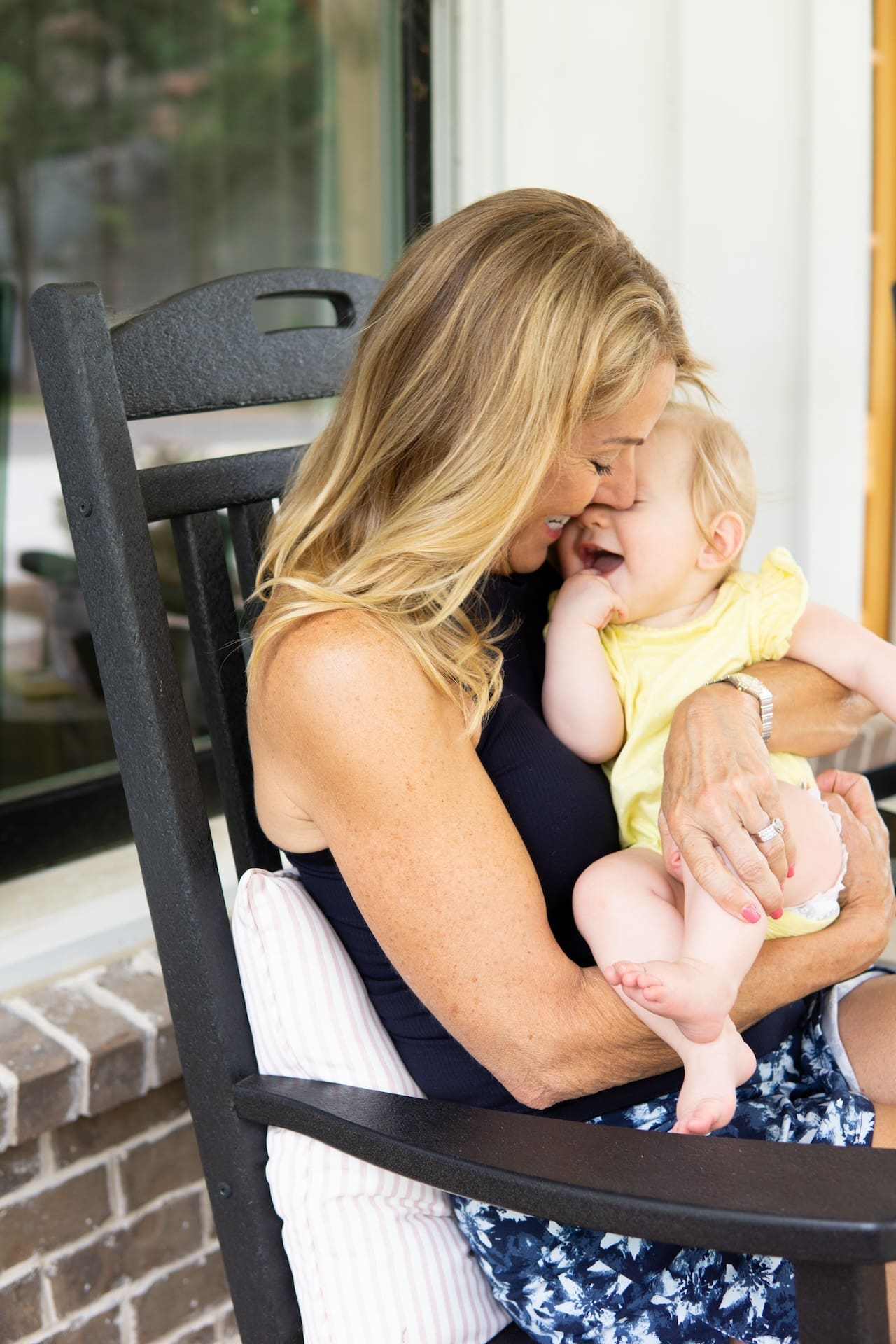 Grandmother cuddles baby in yellow polka dot onesie while sitting in a black rocking chair on a red brick front porch.