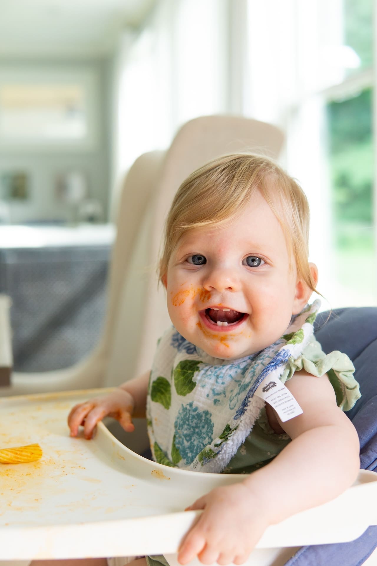 Infant sits in white high chair with hydrangea print bib. Pasta with red sauce on tray and sauce on baby's face.