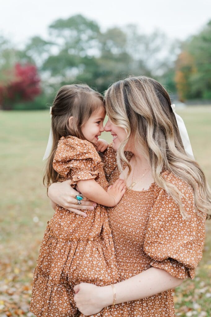 Mother holds toddler daughter in arms as they face one another and nuzzle their noses together. They wear matching orange dresses.