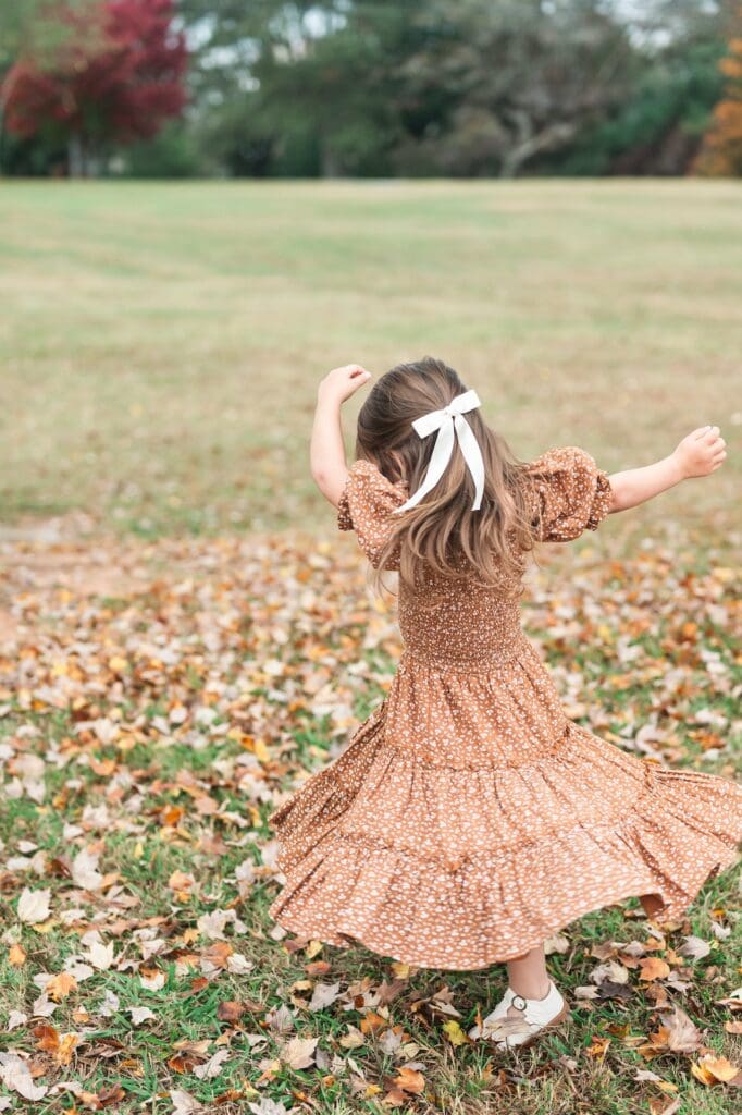 Little girl in orange dress and white bow in her hair dances in field with fall leaves on the ground.