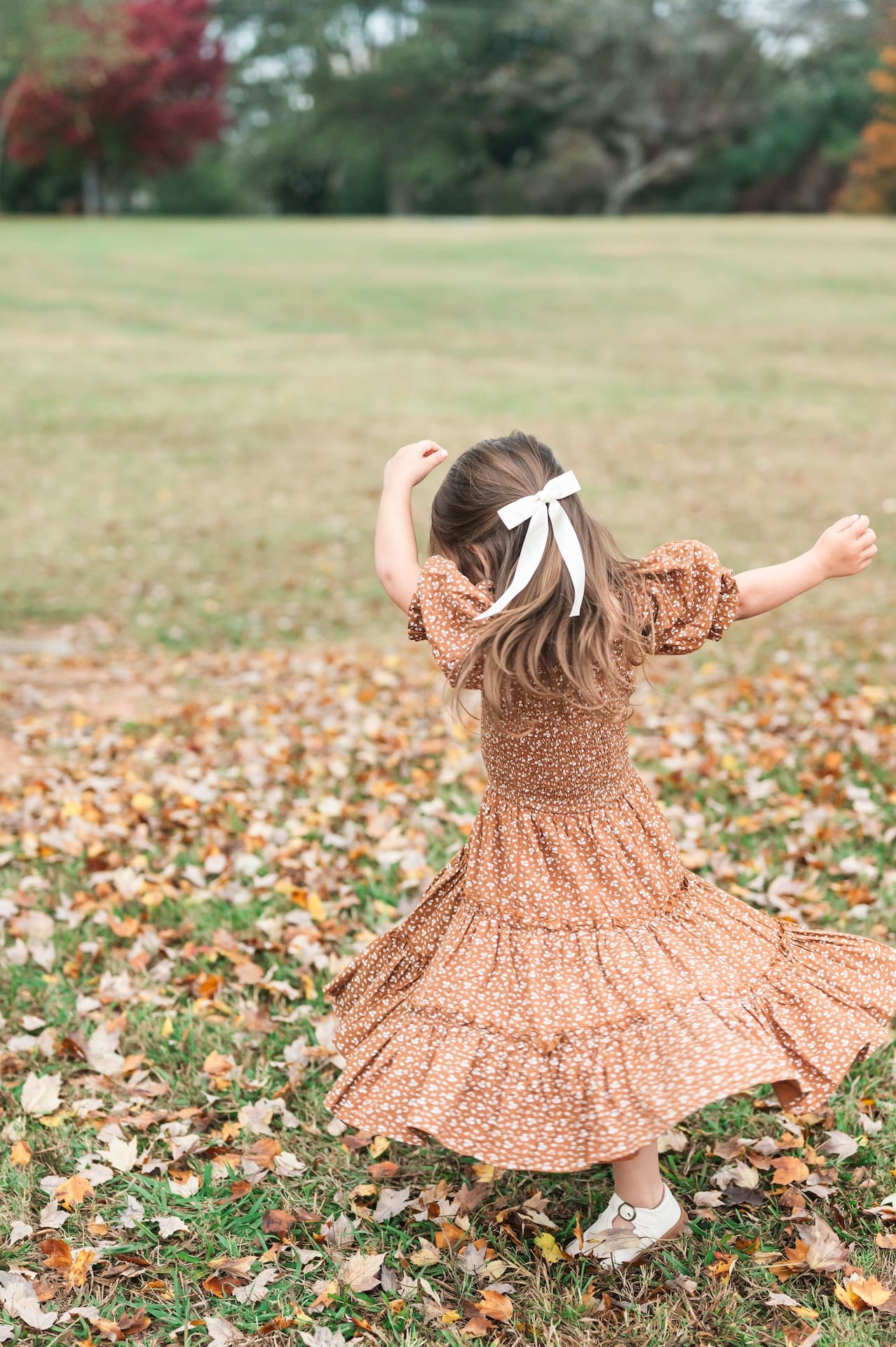 Little girl in orange dress and white bow in her hair dances in field with fall leaves on the ground.
