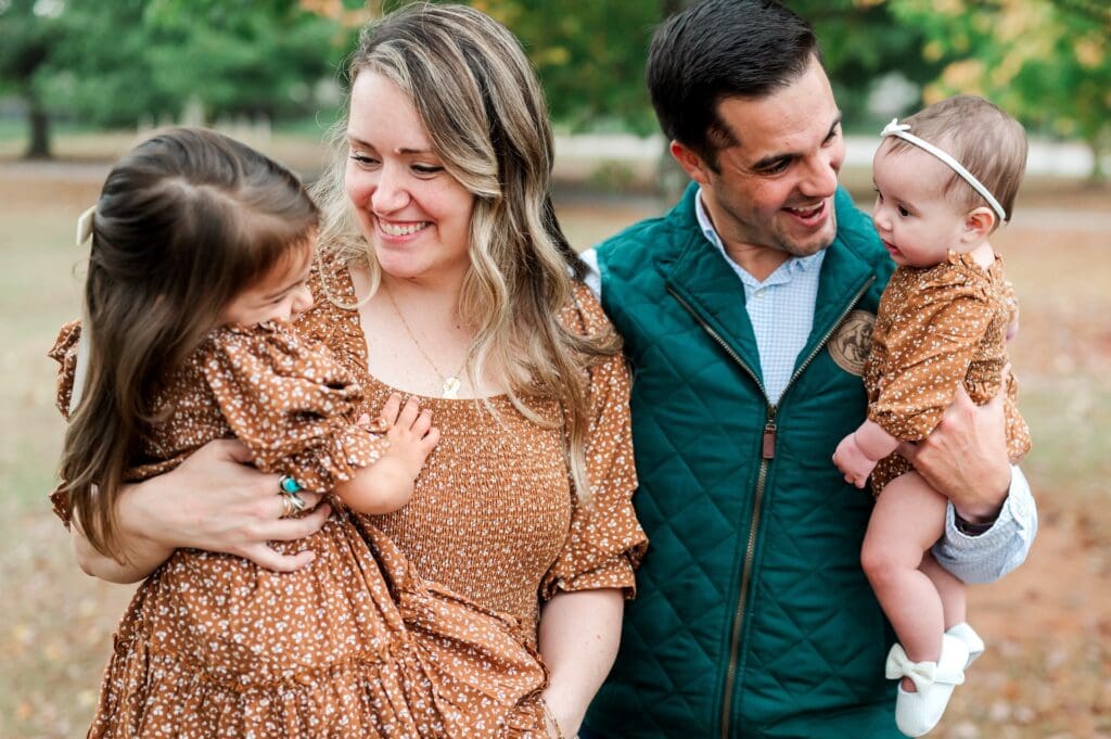 Husband and wife hold daughters in a tree lined field. Mother, toddler and baby are in matching orange dresses.