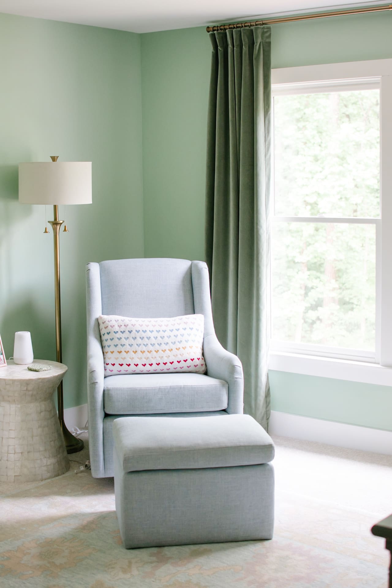 Baby girl nursery with green walls. Image shows blue glider with ottoman, capiz shell side table and gold floor lamp next to a window with green velvet curtains.