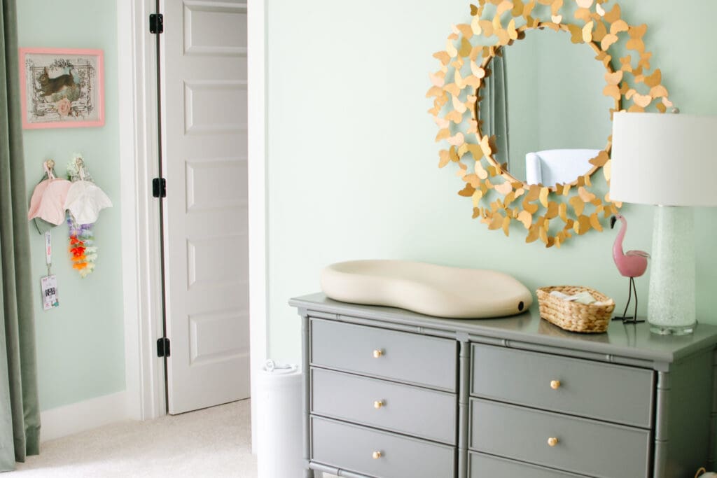 Baby girl nursery with green walls. Image shows grey dresser with butterfly bordered mirror hanging above. Changing pad, water hyacinth basket and green lamp sit on top. Wall to the left has various hats hanging from knobs with a bunny picture in a pink frame above.