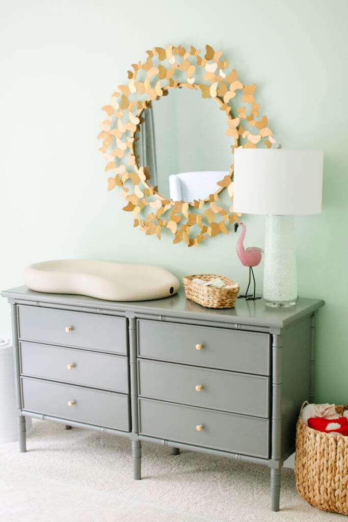 Baby girl nursery with green walls. Image shows grey dresser with butterfly bordered mirror hanging above. Changing pad, water hyacinth basket and green lamp sit on top. Water hyacinth basket with blankets sits on floor next to dresser.