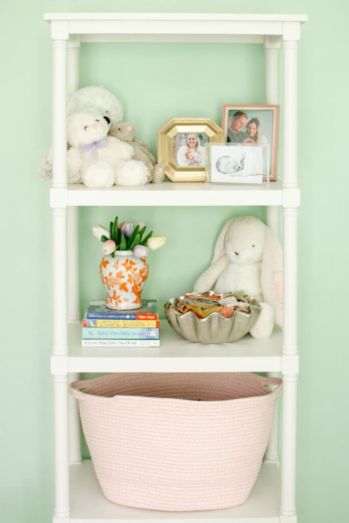 Baby girl nursery with green walls. Image shows white open shelf book case with various family pictures in frames, bunny rabbit stuffed animals, stacked books with an orange chinoiserie vase on top and a pink rope style basket.