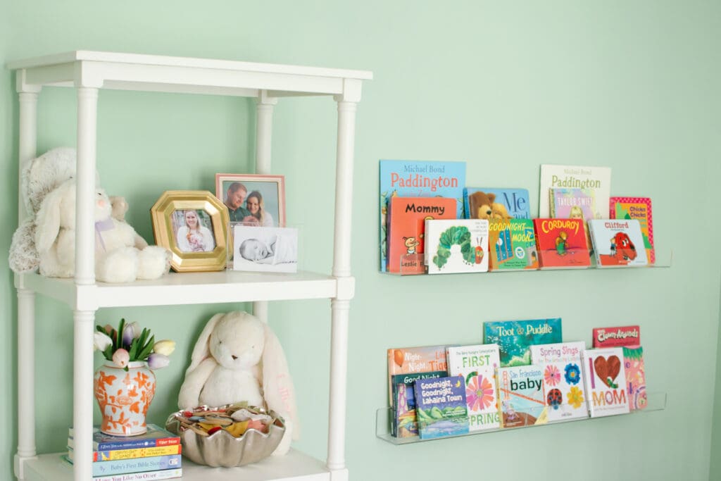 Baby girl nursery with green walls. Image shows white open shelf book case with various family pictures in frames, bunny rabbit stuffed animals, stacked books with an orange chinoiserie vase on top. Next to bookshelf are two acrylic wall shelves featuring various children's books.