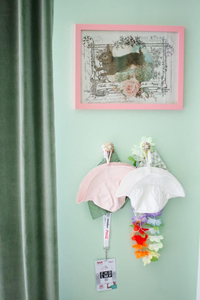 Baby girl nursery with green walls. Image shows close up of wall with hats hanging from knobs with a bunny picture in a pink frame above. Green velvet curtains border the left side of the image.