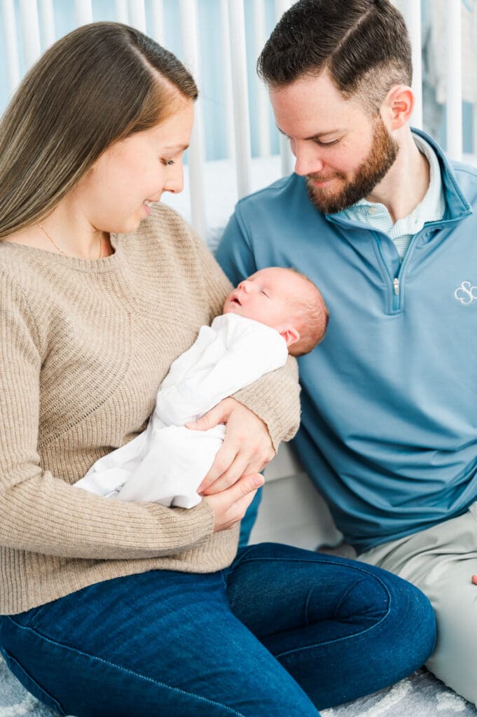 New parents sit leaning against crib staring lovingly at their newborn baby during in-home newborn photography session.