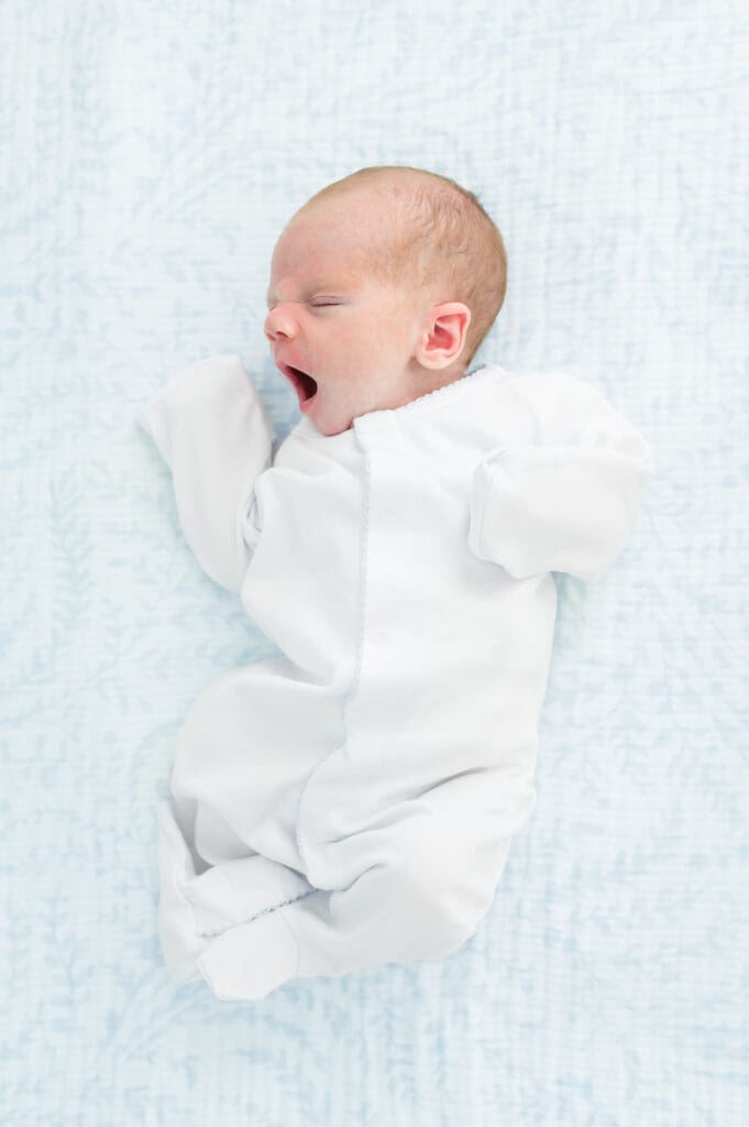 Newborn baby in white onesie lies on back yawning big during in-home newborn photography session.