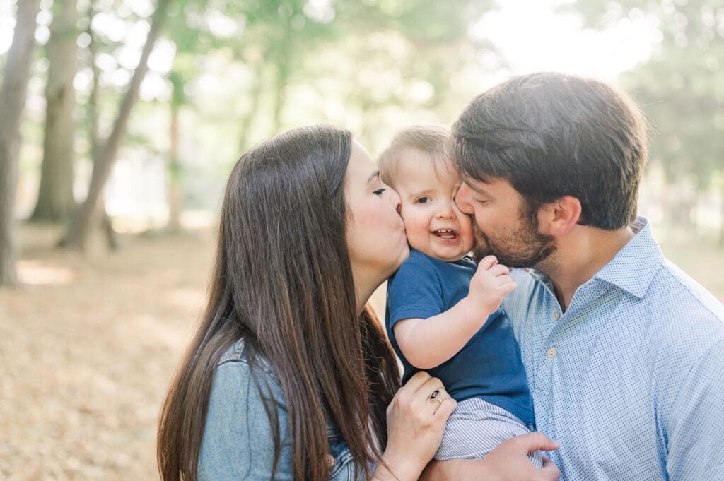 Young couple hold toddler son and kiss both cheeks gently smooshing his face.