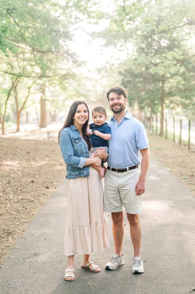 Young couple pose in park while mom holds toddler boy between them. All are smiling.