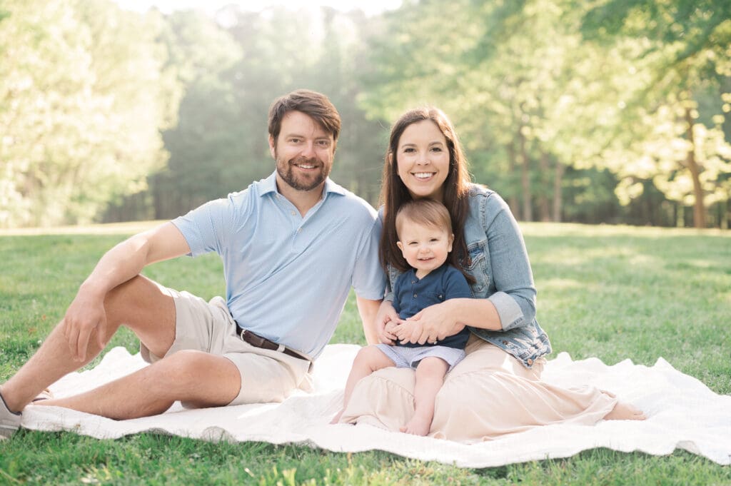 Young mom and dad sit on a white blanket in a park with toddler son on mom's lap. All are smiling.