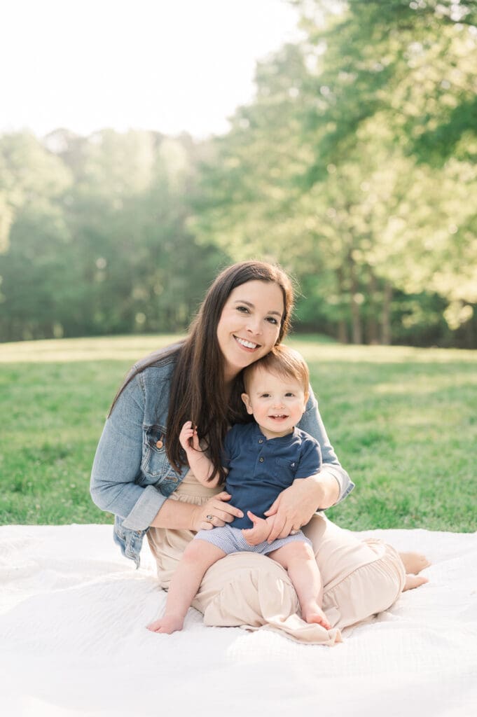 Mom sits on a white blanket in a park with toddler son on her lap. Both are smiling.