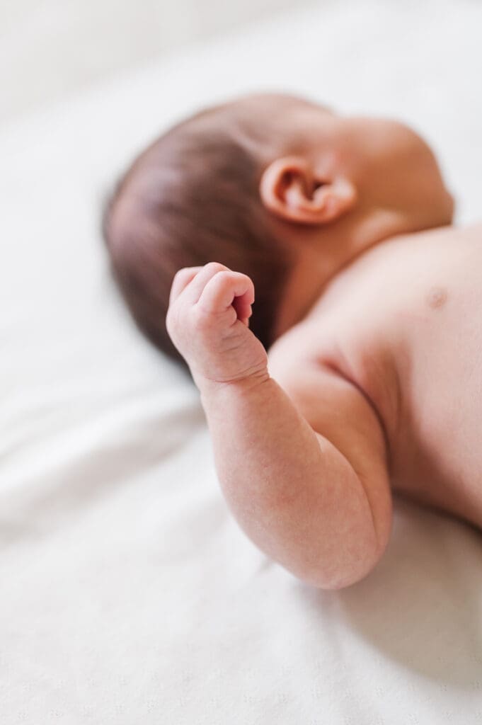 Newborn baby lays on white sheet facing away with a clenched fist.