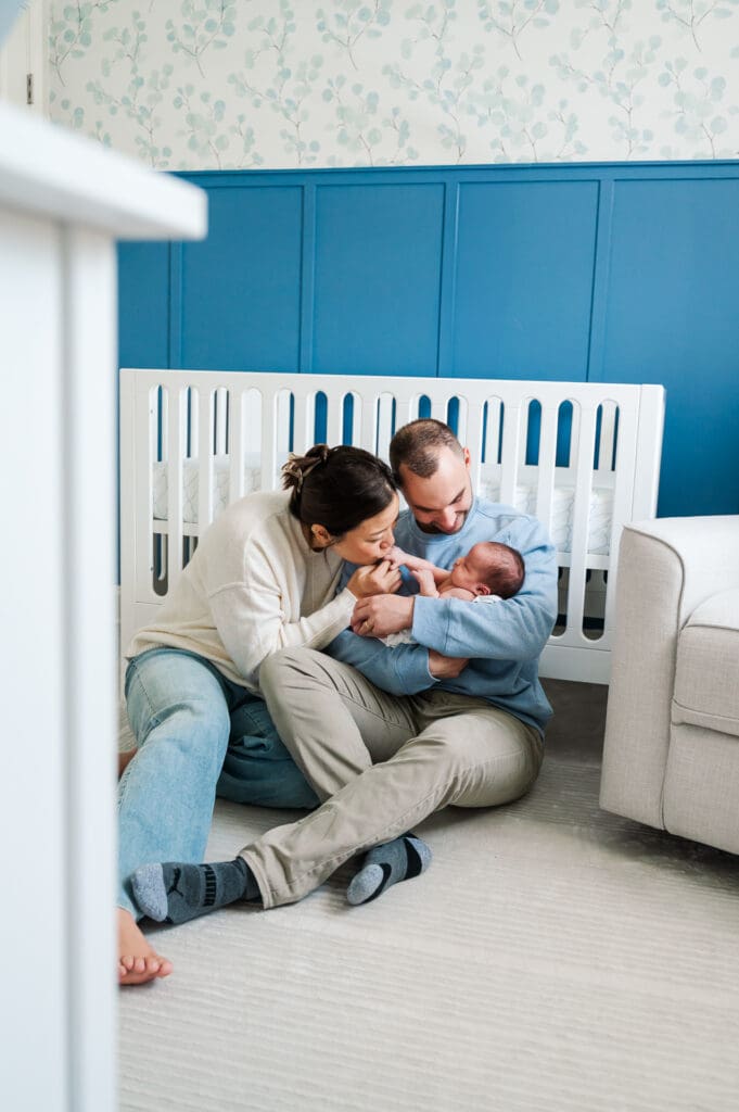 New parents hold new baby as they lean against crib in nursery.