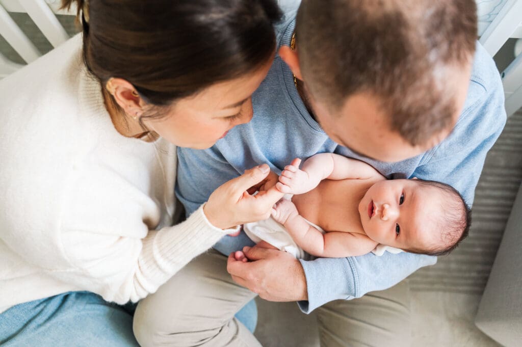 New parents hold new baby as they lean against crib in nursery during newborn photography session.