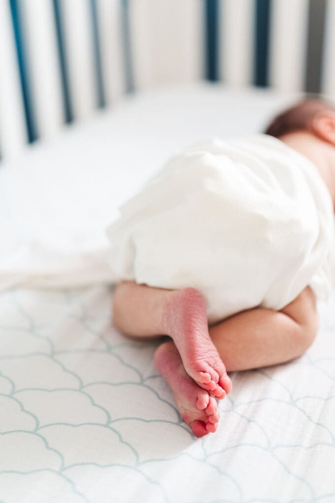 Newborn baby lays on stomach in crib wrapped in white swaddle with feet exposed.