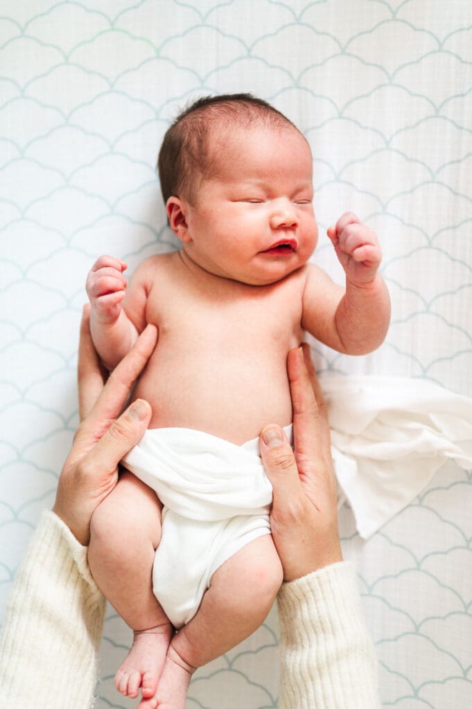 Newborn baby lays in crib wrapped in white swaddle while moms hands wrap around her sides.
