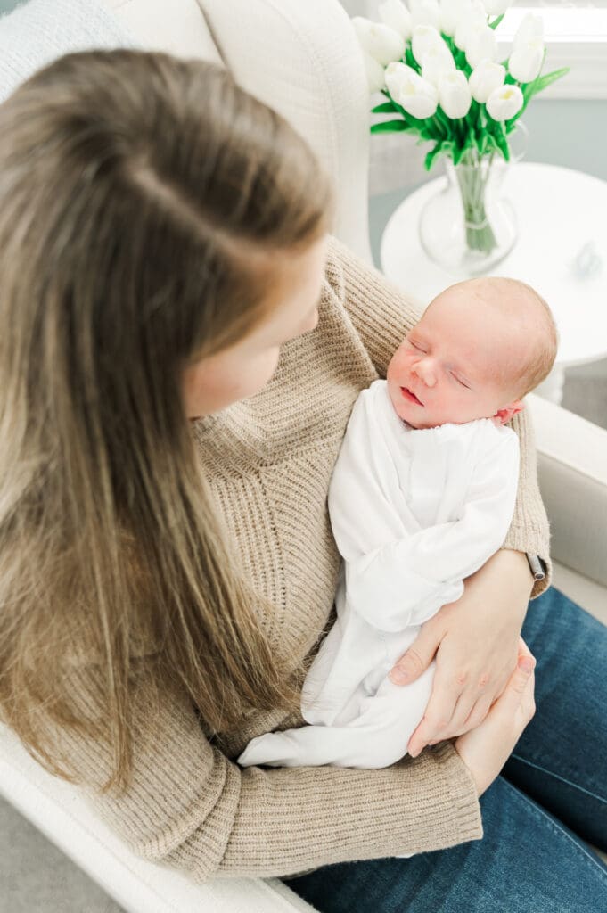 New mom sits in rocking chair holding her newborn baby looking down at him lovingly.