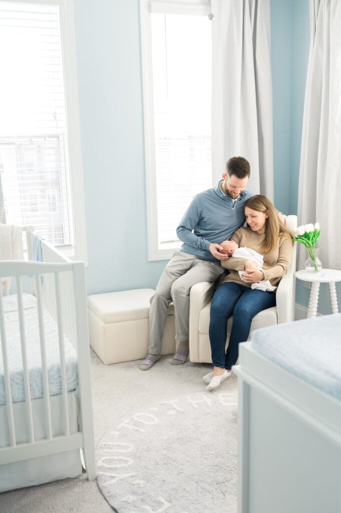 New parents sit in rocking chair in nursery staring lovingly at their newborn baby during in-home newborn photography session.