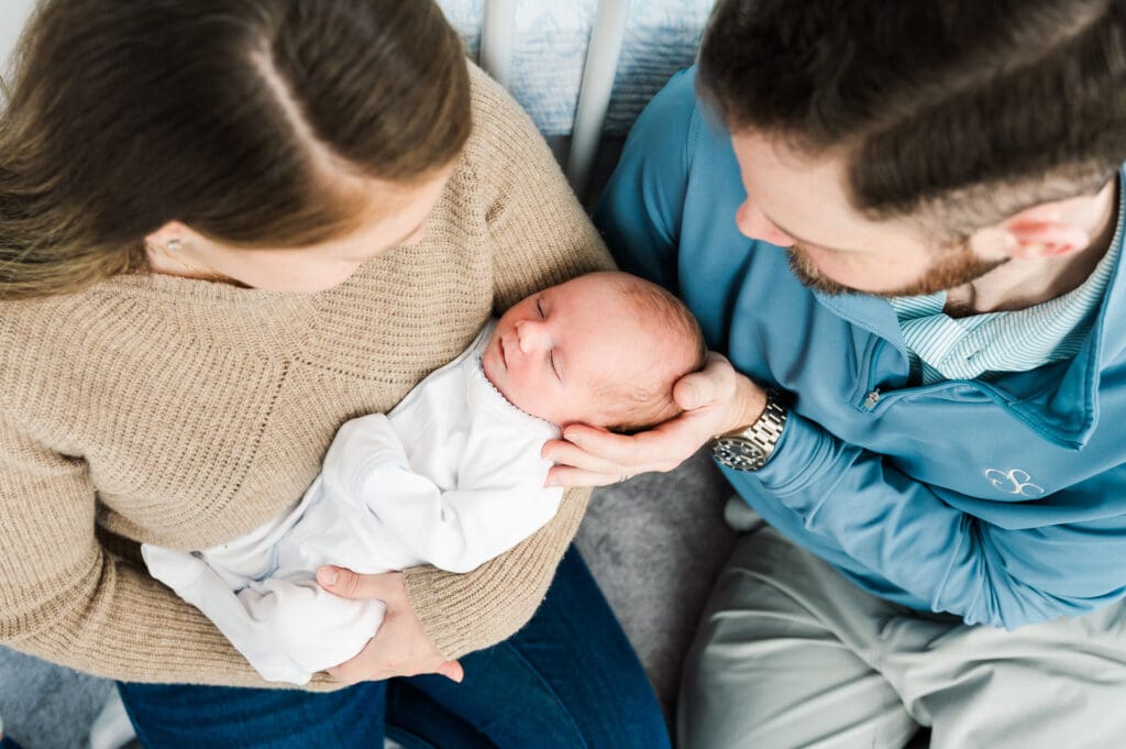 New parents sit leaning against crib staring down lovingly at their newborn baby.