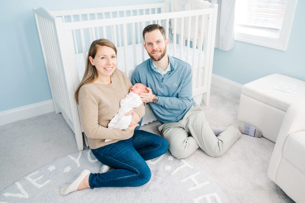 New parents sit leaning against crib smiling as mom holds their newborn baby during in-home newborn photography session.